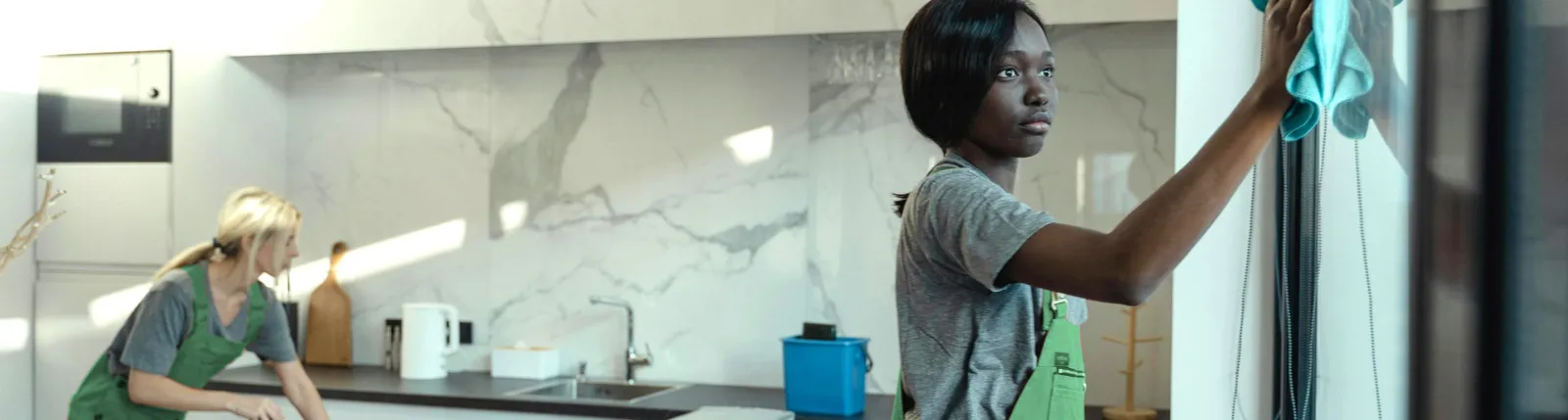Two cleaners working in a bright modern kitchen, one wiping cabinets and one cleaning the counter